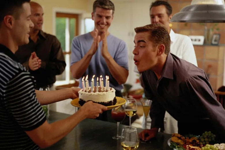 Чоловік дмухає свічки на торт під привітання друзів.
